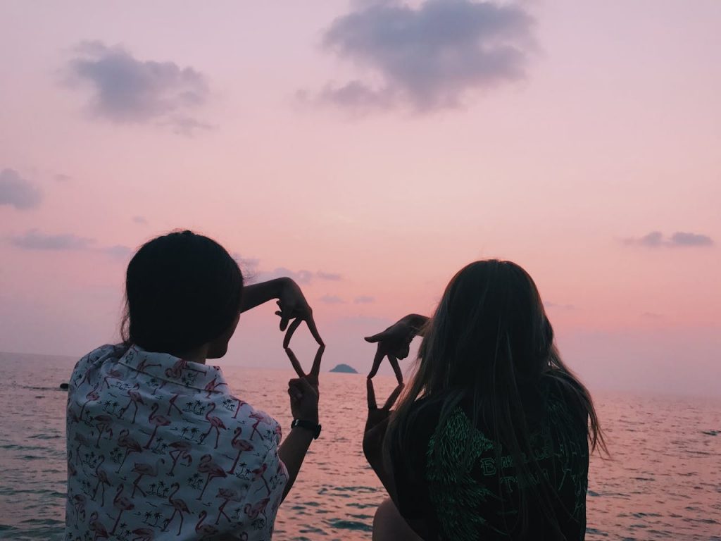 Two friends form heart shapes with their hands by the ocean at sunset, enjoying a peaceful moment.